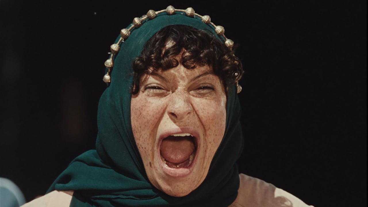Person wearing a dark green headscarf with decorative beads, expressing strong emotion with eyes closed and mouth open.