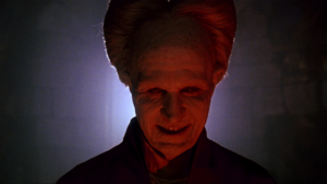 Person with exaggerated high-piled hair and a sinister smile, lit from below with red light and a blue light in the background.