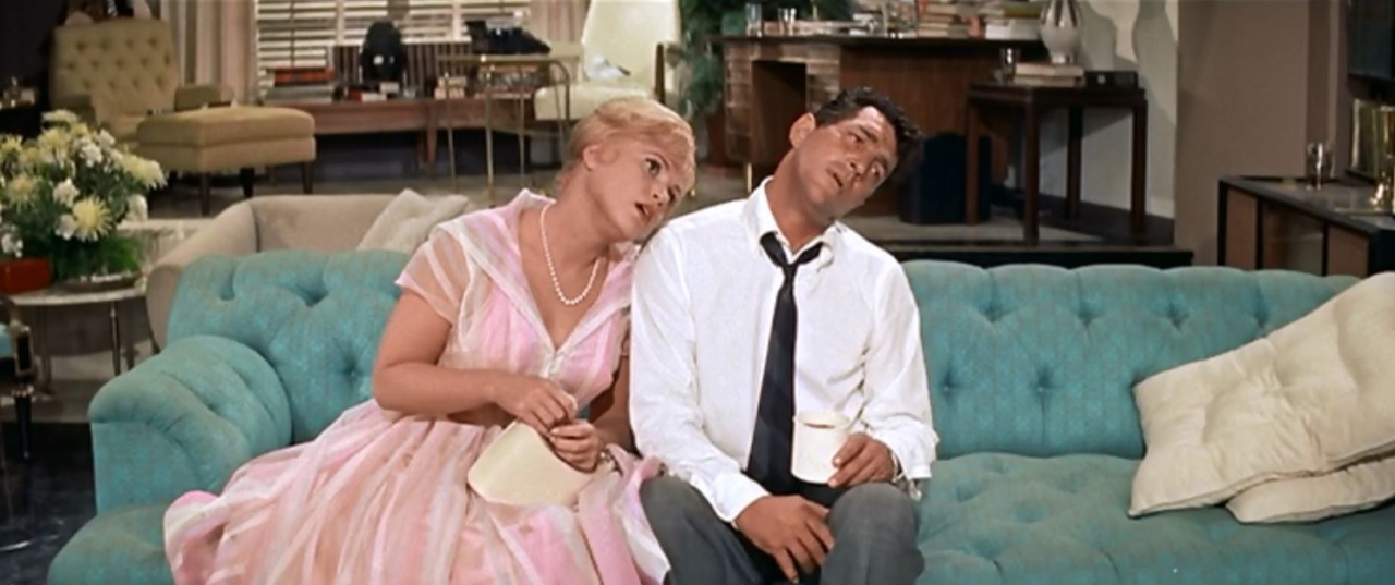 A woman in a pink dress and a man in a shirt and tie sit on a turquoise sofa, leaning toward each other. The room is stylishly decorated with furniture and flowers.