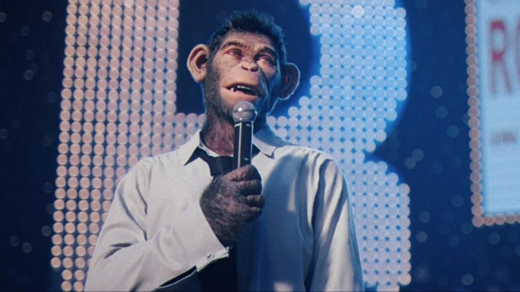 A chimpanzee in a white shirt and tie holds a microphone on stage, with a backdrop of illuminated dots.