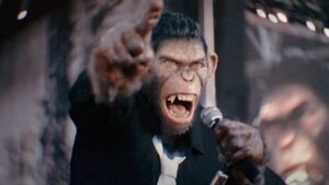 A chimpanzee in a suit holds a microphone and points forward, appearing to be speaking or performing on a stage.