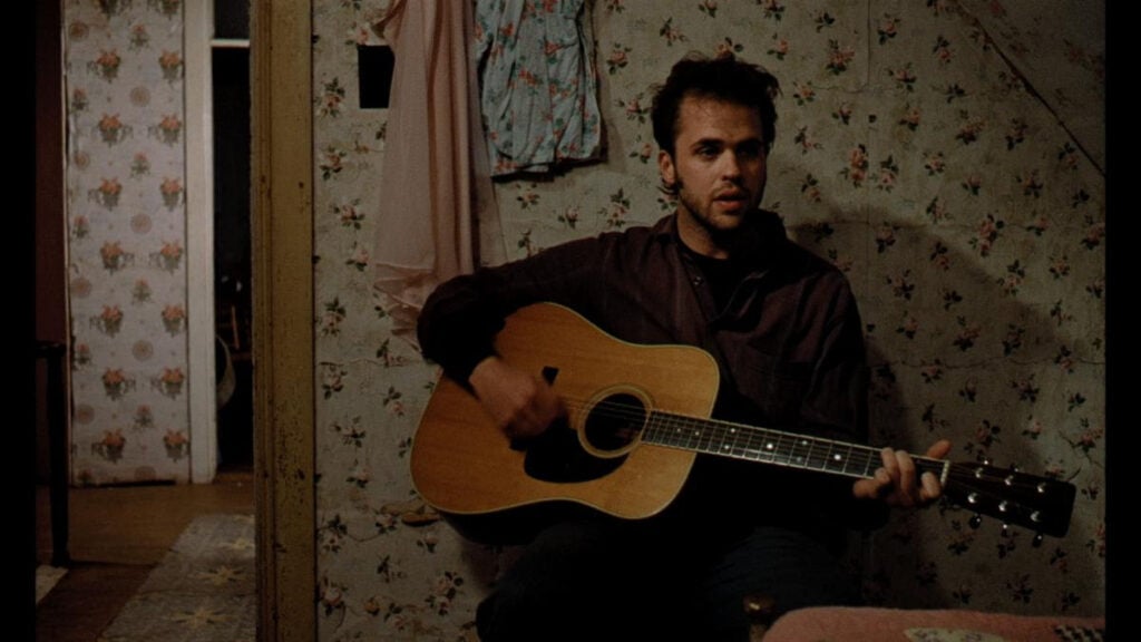 A man sits on a bed, playing an acoustic guitar in a dimly lit room with floral wallpaper and clothing hanging on the wall.