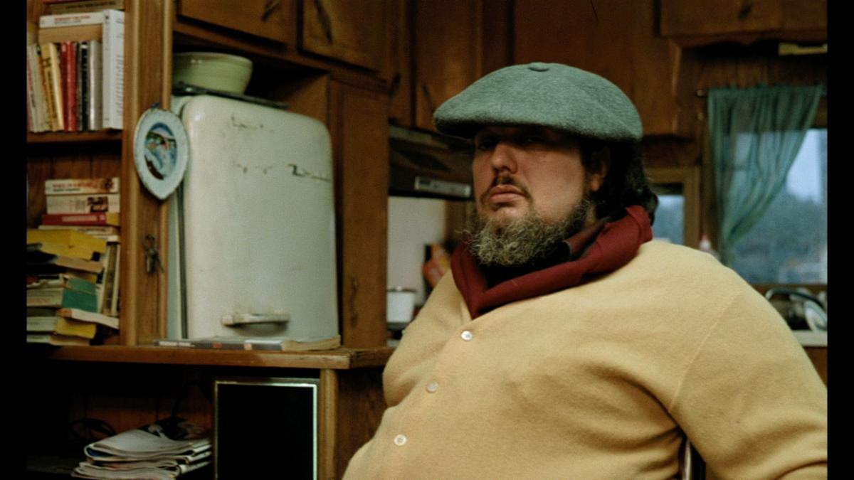 A man wearing a gray flat cap and a yellow cardigan sits in a kitchen with wooden cabinets and an old refrigerator.