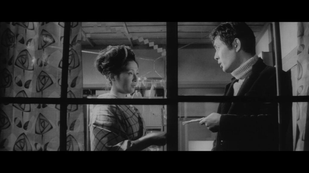 A man and woman converse through a window. The woman wears a patterned robe and the man a dark jacket and turtleneck. The room has a patterned curtain and visible ceiling beams.