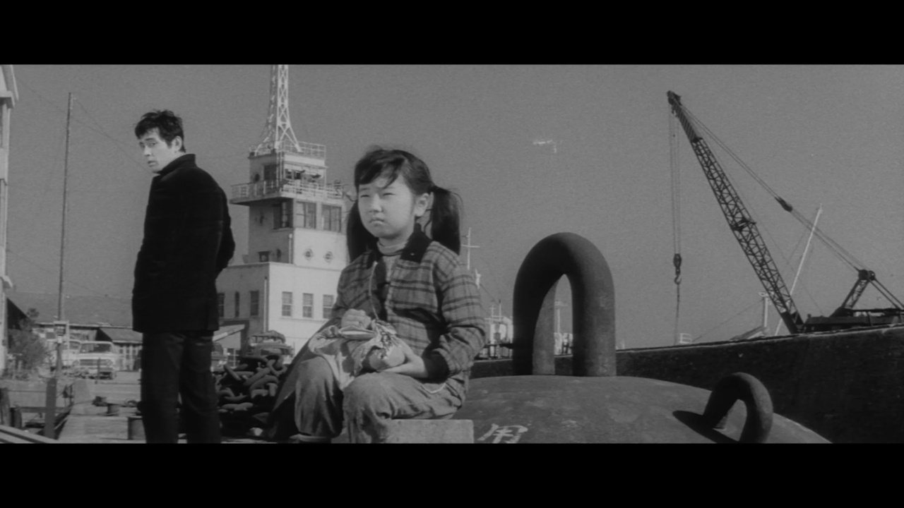 A girl sits on a large object, holding a stuffed animal, while a man stands in the background. An industrial area with a tower and cranes is visible under a clear sky.