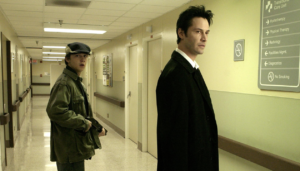 Two men stand in a hospital hallway; one wears a coat and holds a gun, the other wears a suit. Signs indicating various departments are visible on the wall.