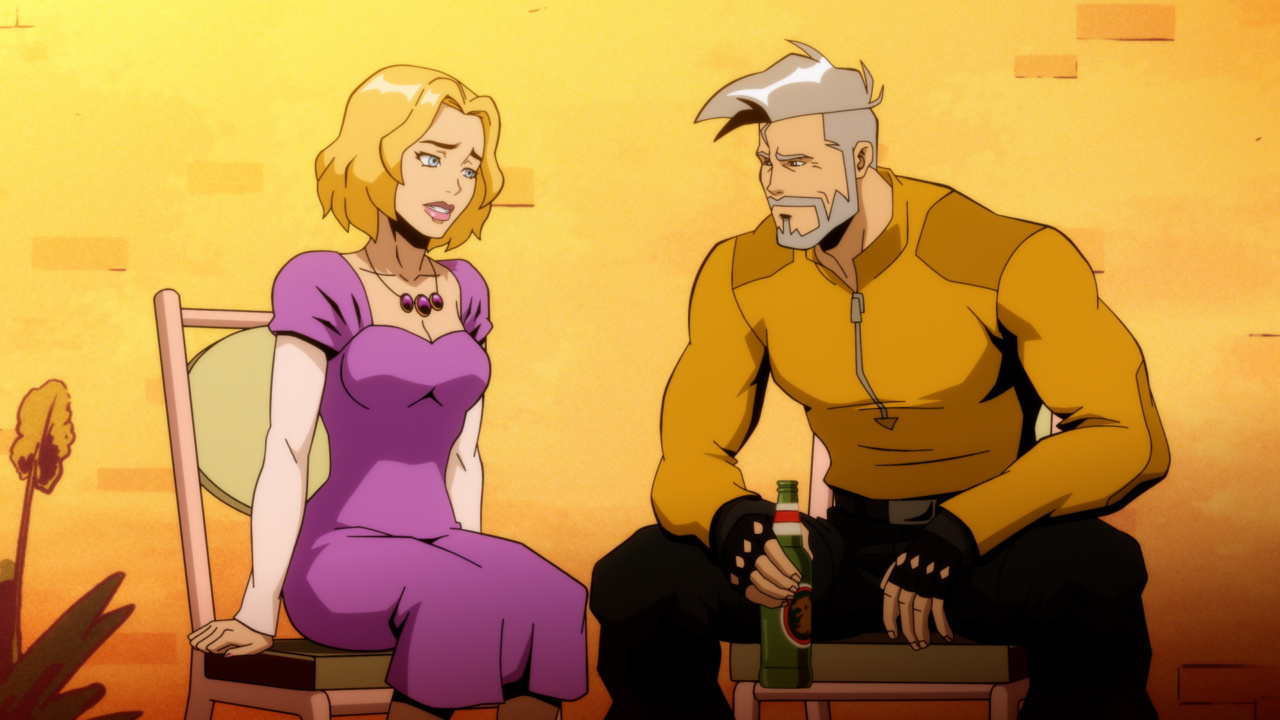 A woman in a purple dress sits next to a muscular man with gray hair, wearing an orange shirt and holding a bottle, both sitting on chairs against an orange backdrop.