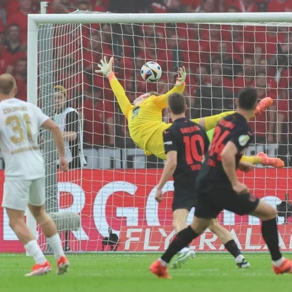 A goalkeeper in yellow makes a leaping save against a team in black jerseys during a soccer match, with a white-jerseyed player in the foreground and spectators in the background.