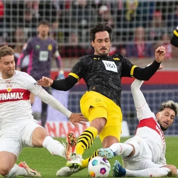 Three soccer players from opposing teams compete for the ball on a field, with one player in black and yellow kit falling to the ground. Other players in white and red attempt to tackle the ball.