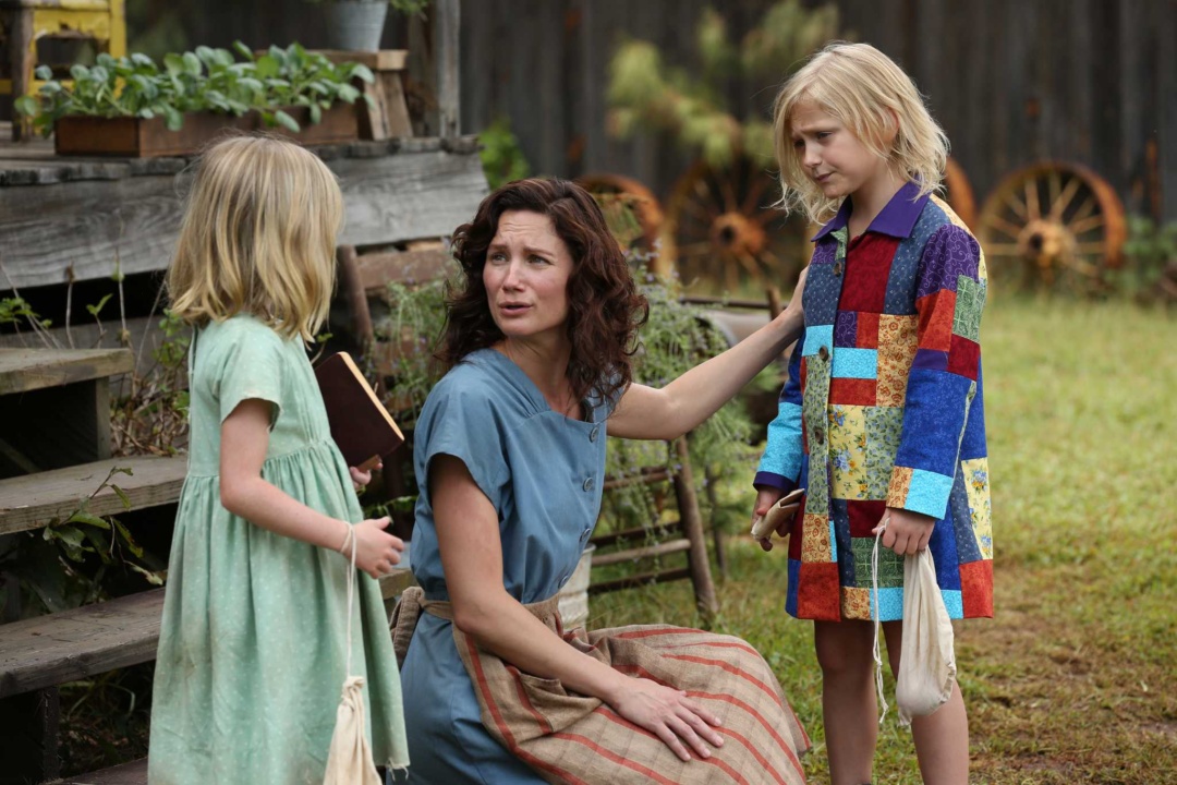 A woman kneels between two children in colorful dresses, outdoors near wooden steps. The woman is talking to them, one child holds a book, the other a bag.