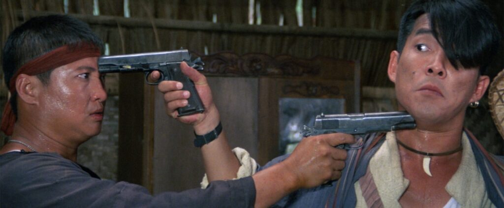 Two men stand facing each other indoors, holding guns to each other's heads. Both appear tense, with one man wearing a headband and the other sporting a distinctive haircut.