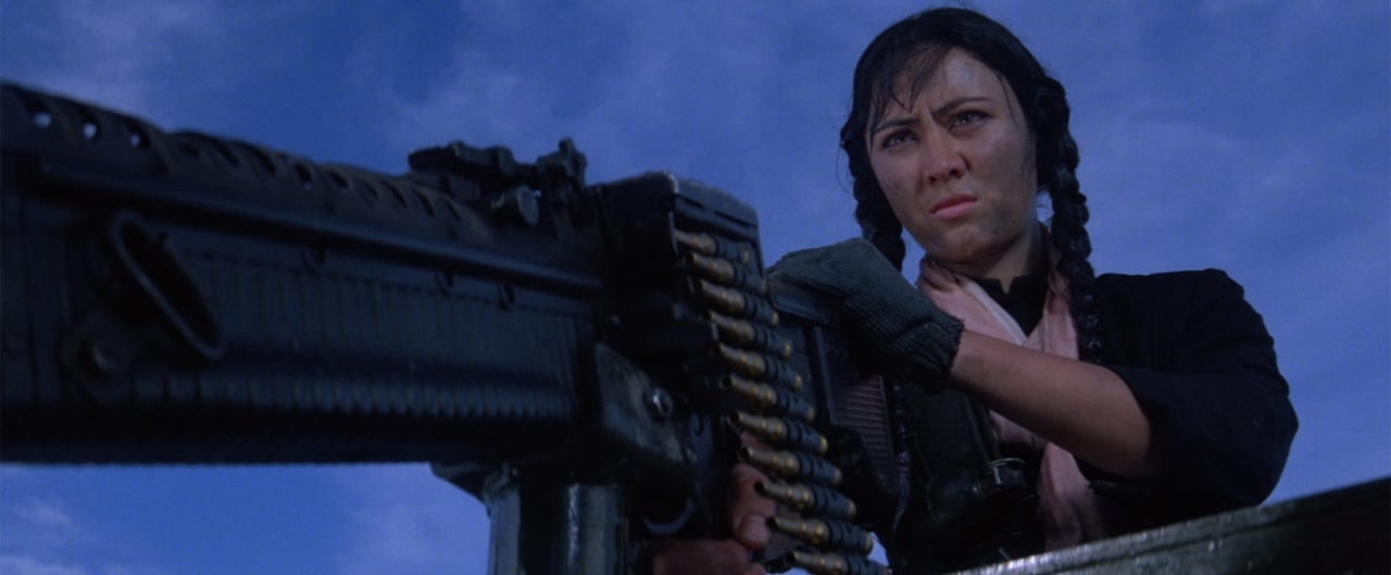 Person with braided hair operates a large mounted machine gun against a blue sky backdrop.