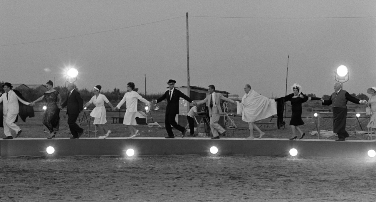A group of people in various outfits is walking in a line on a lit platform outdoors, during the evening.