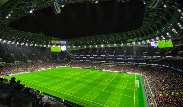 A large, illuminated football stadium is filled with spectators watching a match, with players on the field and vibrant green grass.