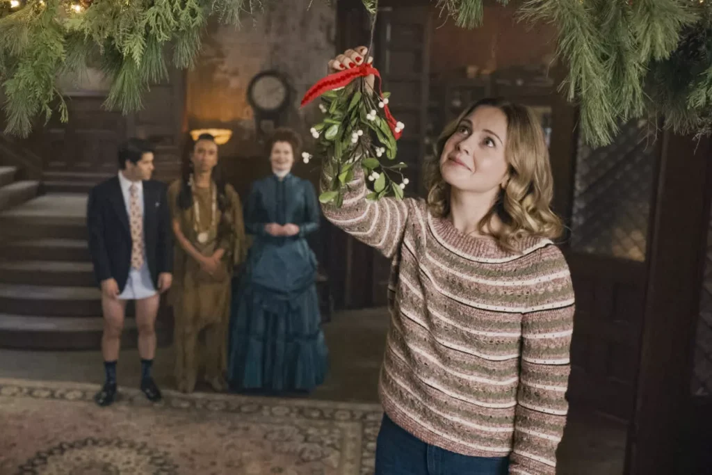 A woman in a sweater hangs a mistletoe indoors, while three people, including one in a blue dress and another without pants, watch in the background.