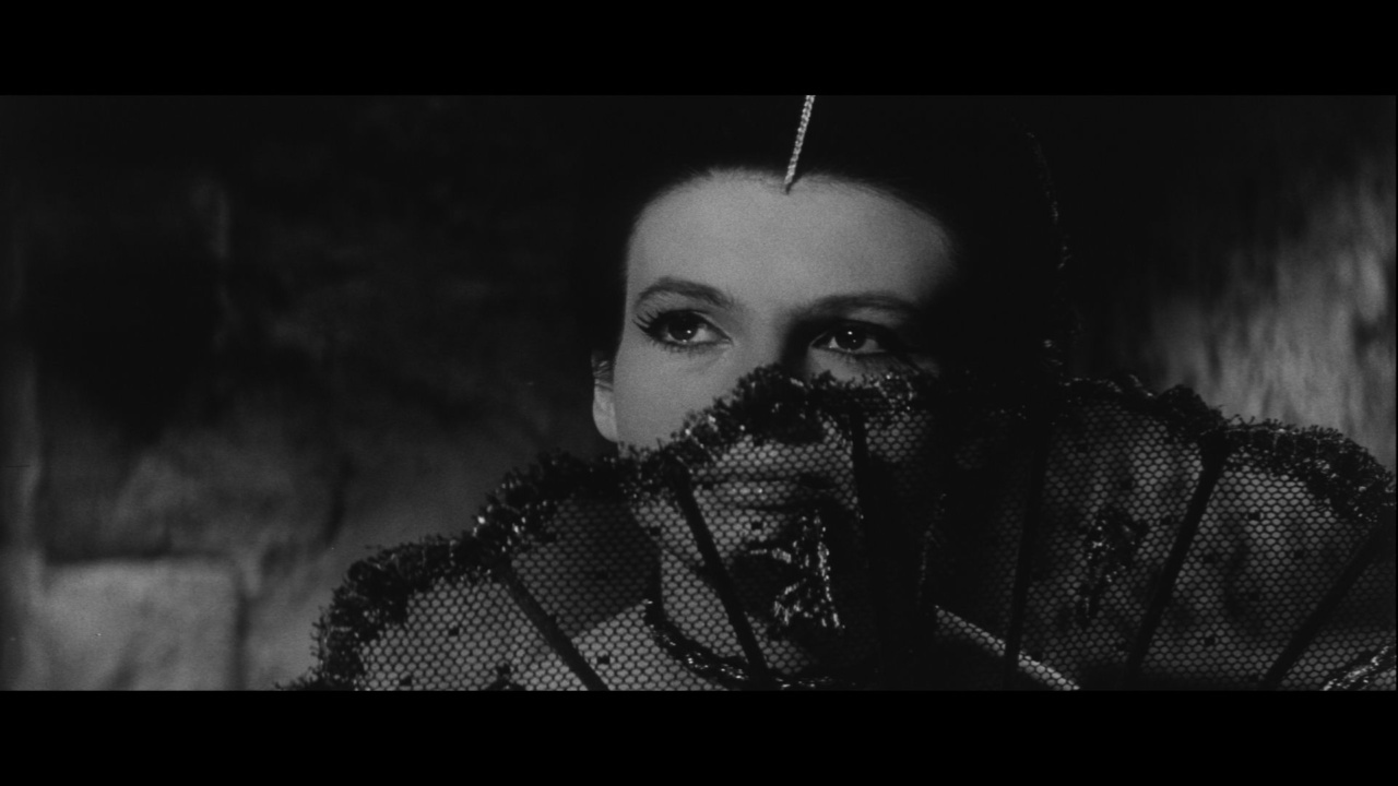A woman partially covers her face with a lace fan, gazing intently to the side in a dimly lit setting.