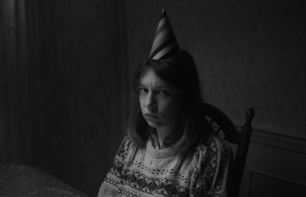 A person with a party hat sits at a table, wearing a sweater, looking displeased. The image is in black and white.