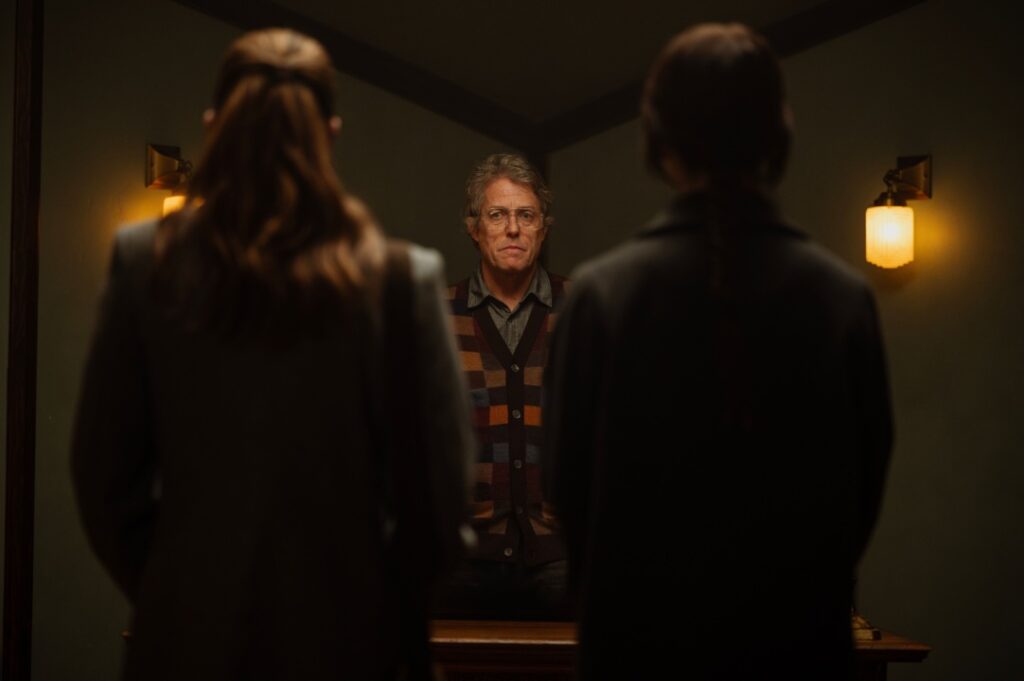 A man in glasses and a colorful sweater stands in a dimly lit room, facing two people whose backs are to the camera. Two wall sconces provide subtle lighting.
