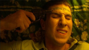 A person with "USDA PRIME" tattooed on their forehead holds a gun to their head while lying on a floral blanket, appearing tense.