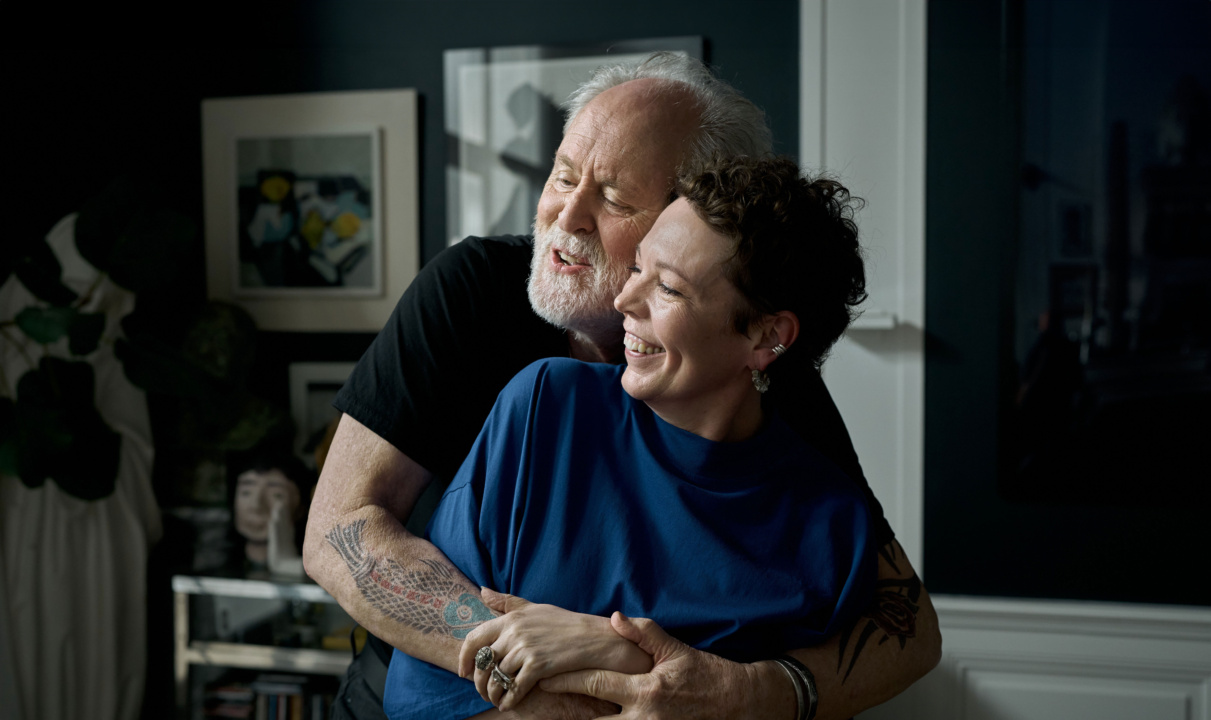 An elderly man with a white beard hugs a smiling person with short hair and a blue shirt. They stand in a room with art on the walls.