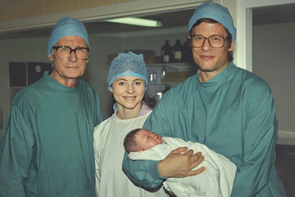 Three medical professionals in surgical attire stand in a hospital room. One person holds a newborn wrapped in a white blanket.