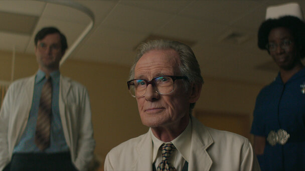 An older man in glasses and a beige coat is seated, with two people in medical attire standing behind him in a room with fluorescent lighting.
