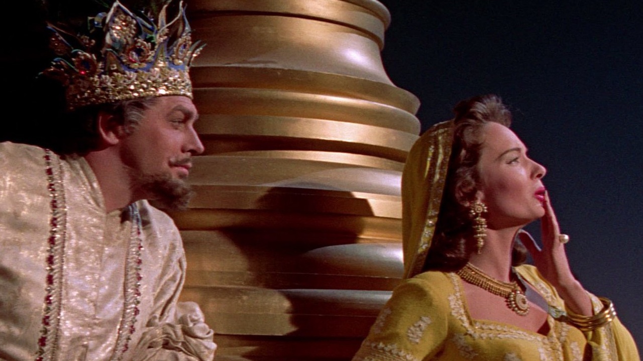 A man wearing a crown and ornate robe stands next to a woman in a yellow dress and headdress, in front of a large golden column.