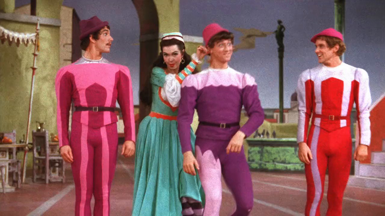 Four people in colorful theatrical costumes stand in a row, with a vibrant background reminiscent of a stage set.