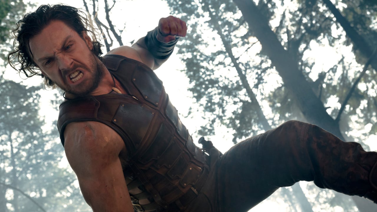 A man in a leather vest with long hair is mid-action, appearing to leap or attack, in a forest setting.