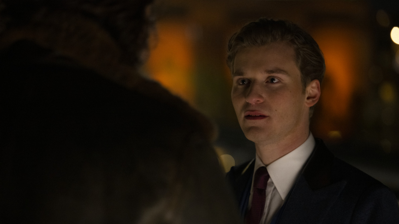 A person in formal attire appears concerned while speaking to someone whose back is turned, in a dimly lit setting.