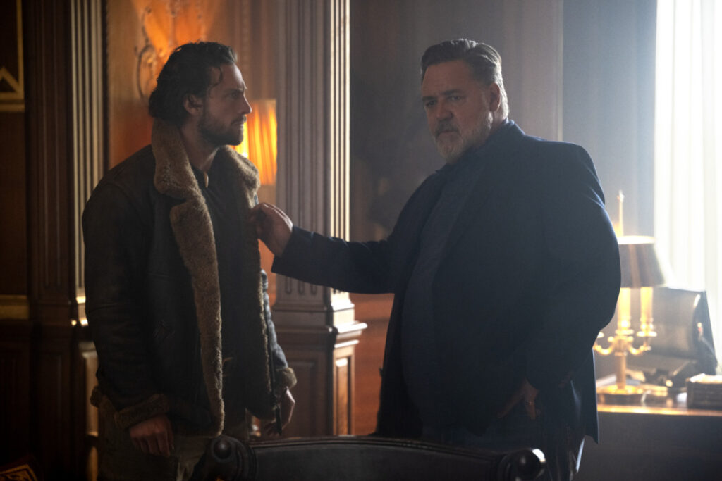Two men stand in a dimly lit room, one in a fur-lined coat and the other in a blazer, engaged in a serious conversation.