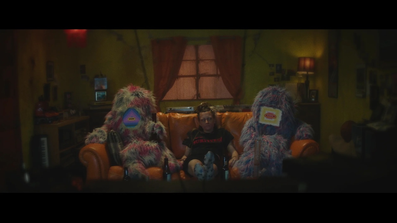 A person sitting on a couch between two fuzzy, colorful creatures in a dimly lit room with yellow walls and a window in the background.