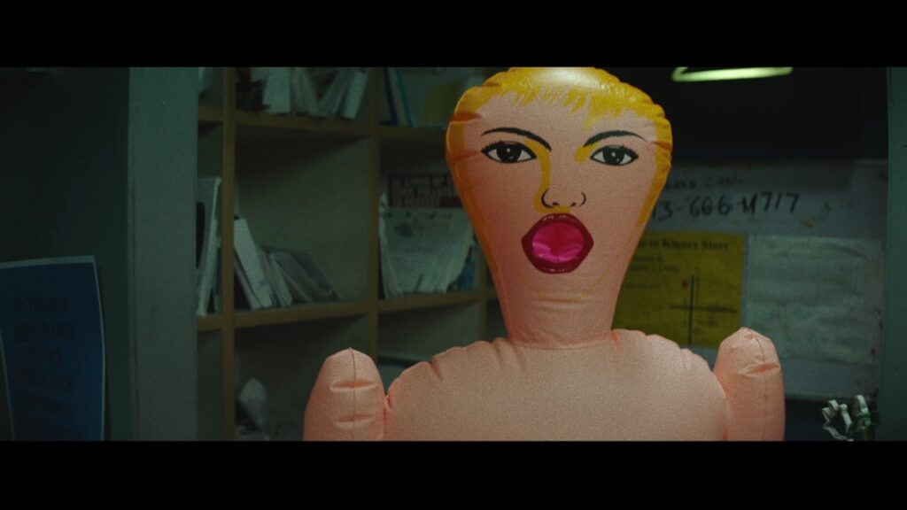 Inflatable doll with facial features stands in a dimly lit room with shelves and posters on the walls.