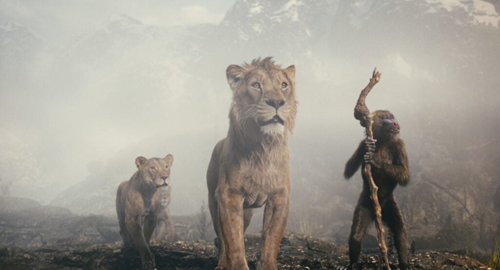 Three animals stand on rocky terrain: a lion, a lioness, and a baboon holding a stick, with foggy mountains in the background.