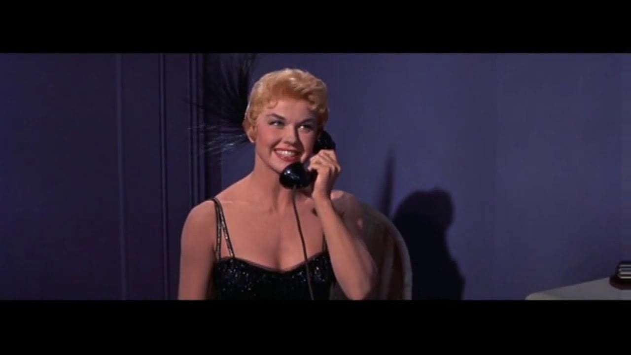 A person with short blonde hair is smiling and talking on a black rotary phone, wearing a black dress, against a dark background.