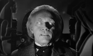 Eureka Entertainment Announces Upcoming Blu-Ray Titles From Kinji Fukasaku, Fritz Lang & More An older man with white eyebrows sits in a chair, eyes rolled back. The background features abstract shapes in black and white.
