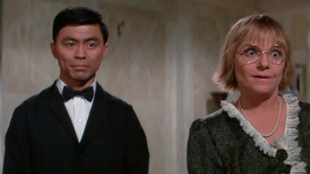 A man in a dark suit and bow tie stands beside a woman with glasses and a green dress in a room with beige walls.