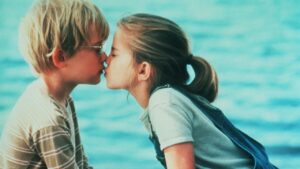 A young boy and girl are kissing in front of a blurred, blue-toned background.