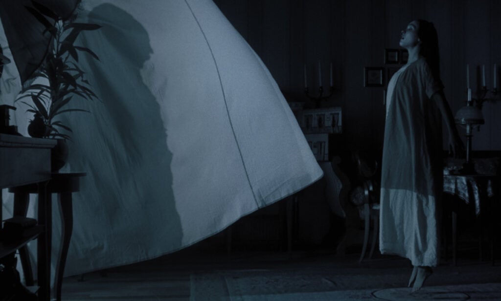 A woman in a white gown levitates in a dimly lit room, with a large billowing sheet nearby.