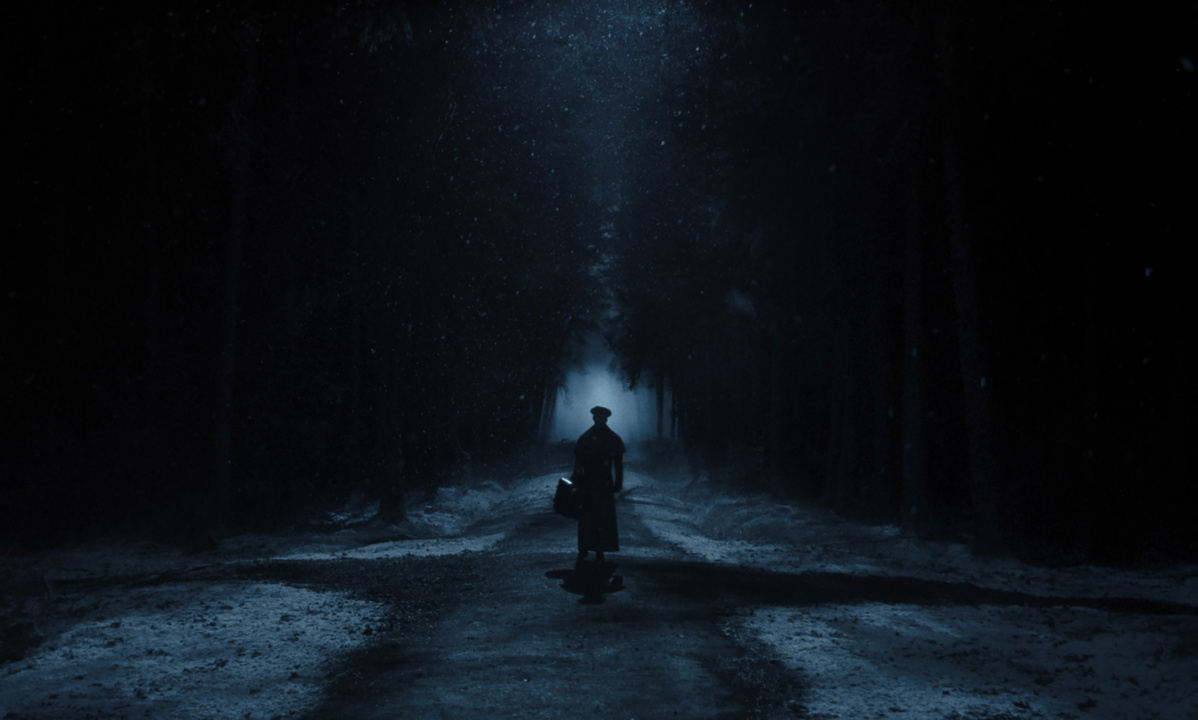 A person in a coat and hat walks down a snowy, moonlit path surrounded by trees, carrying a suitcase.