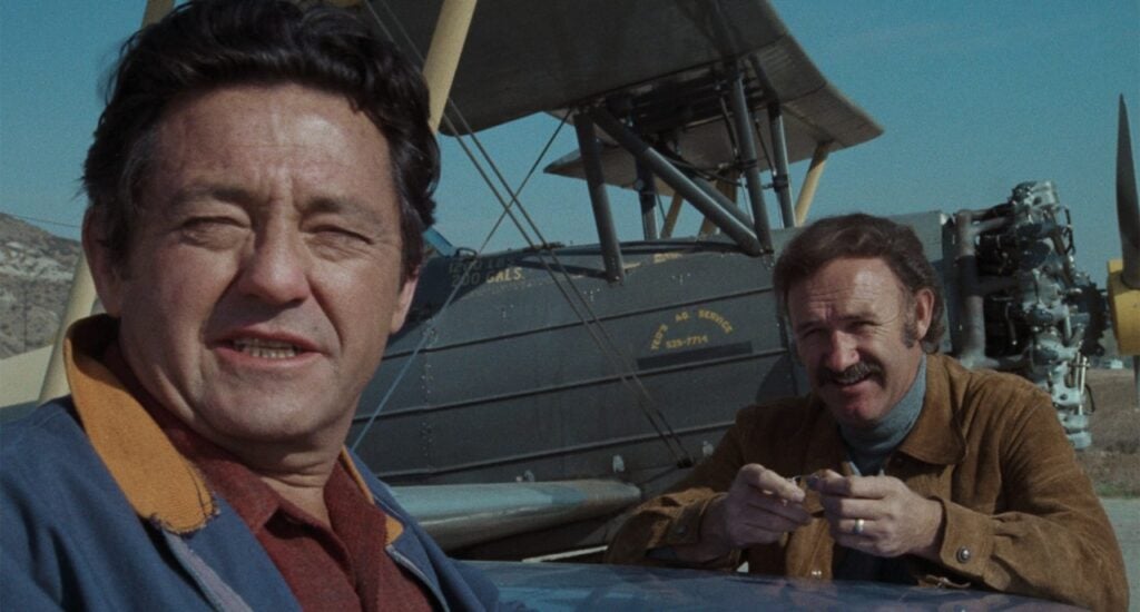 Two men stand in front of a vintage biplane, one leaning forward and the other standing beside the wing.