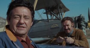 Two men stand in front of a vintage biplane, one leaning forward and the other standing beside the wing.