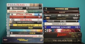 A stack of various DVD and Blu-ray cases against a teal background, featuring titles like "Paris, Texas" and "Joker: Folie à Deux.