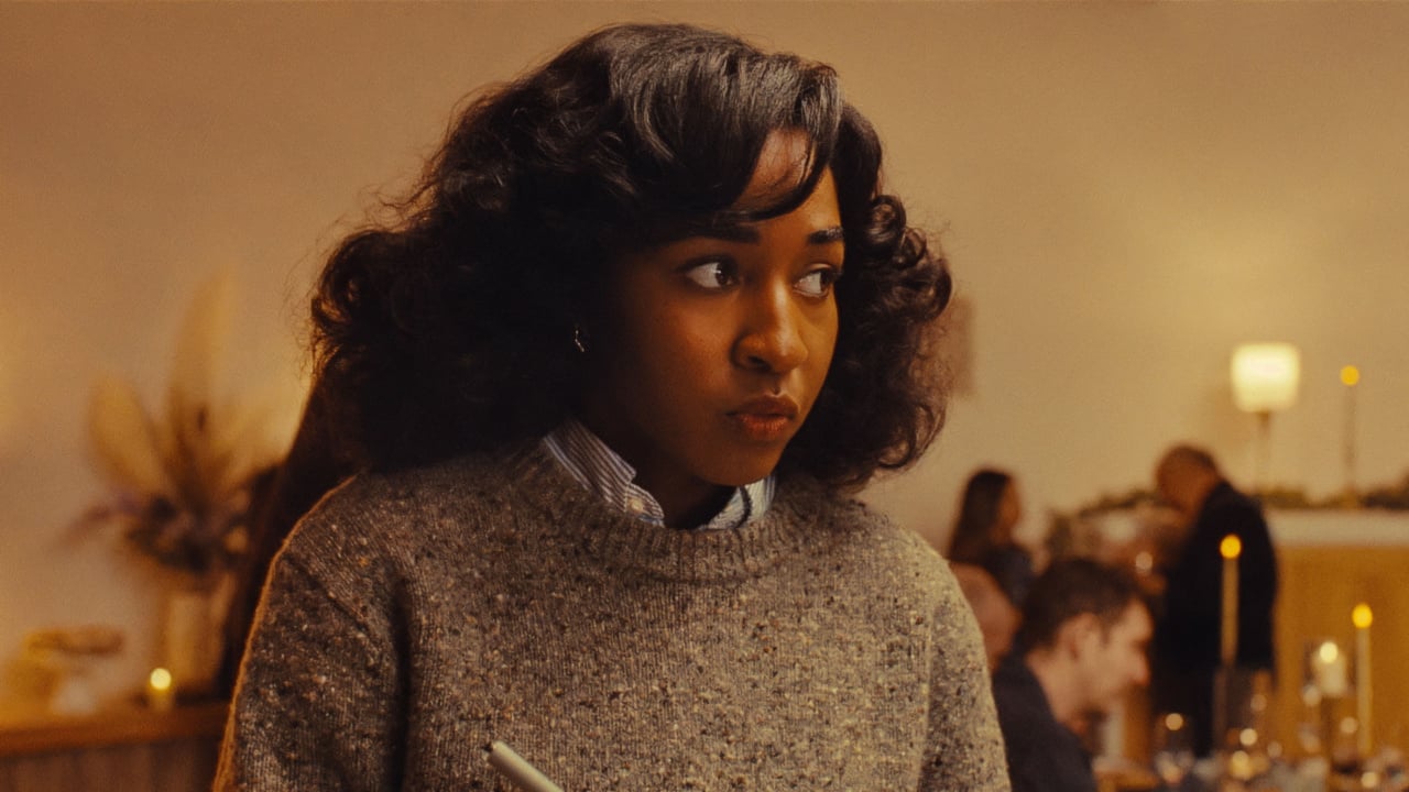 A person with curly hair wearing a sweater is holding a pen, looking off to the side in a warmly lit room.