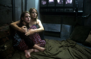 Two people sitting on the floor in a dimly lit room, one holding the other. Monitors are visible in the background.