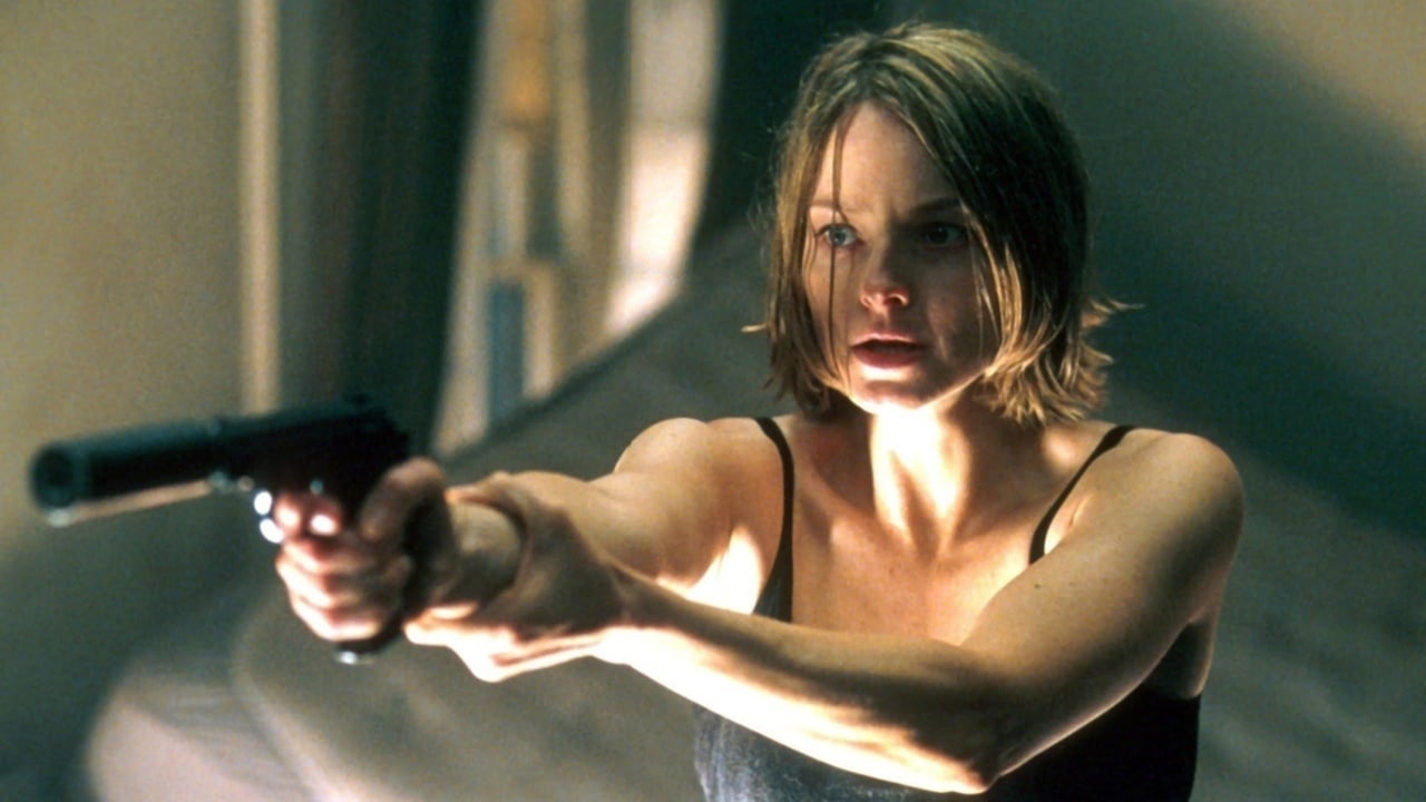 A woman with short hair, wearing a black tank top, is aiming a handgun intently indoors.