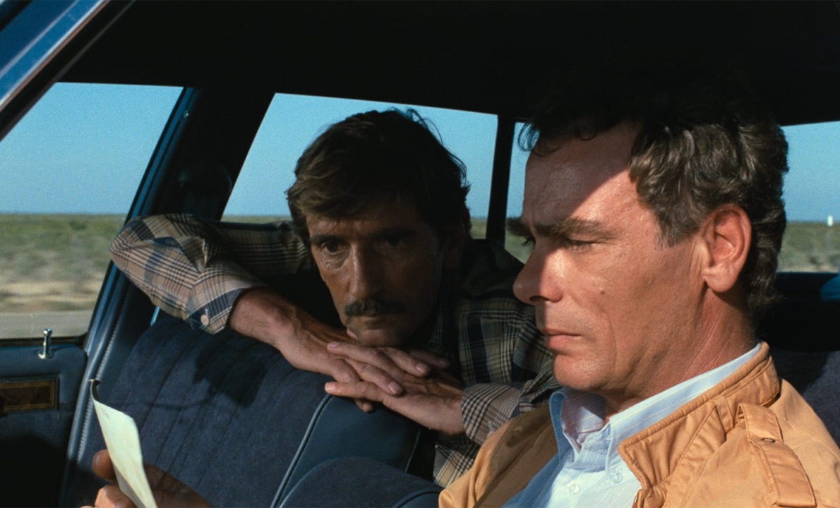 Two men are sitting in a car. One man is looking at a piece of paper while driving, and the other is leaning over from the back seat, watching him. The background shows a desert landscape.