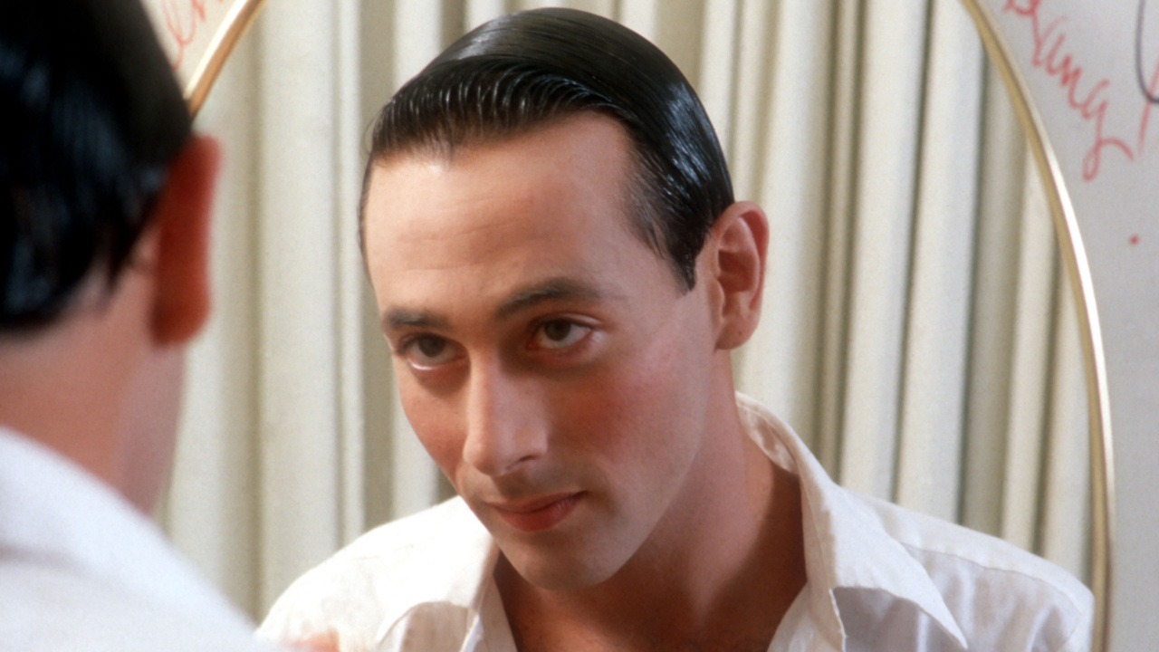 A person with slicked-back hair and a white shirt looks at themselves in a mirror, with writing visible on the glass.