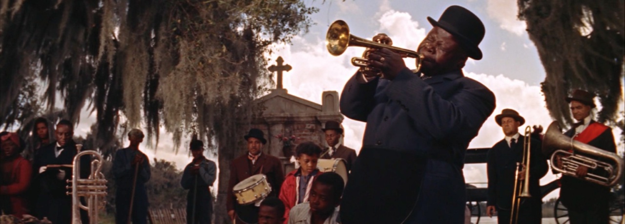 A musician plays the trumpet in a cemetery while a group of onlookers, including other musicians, gather around.