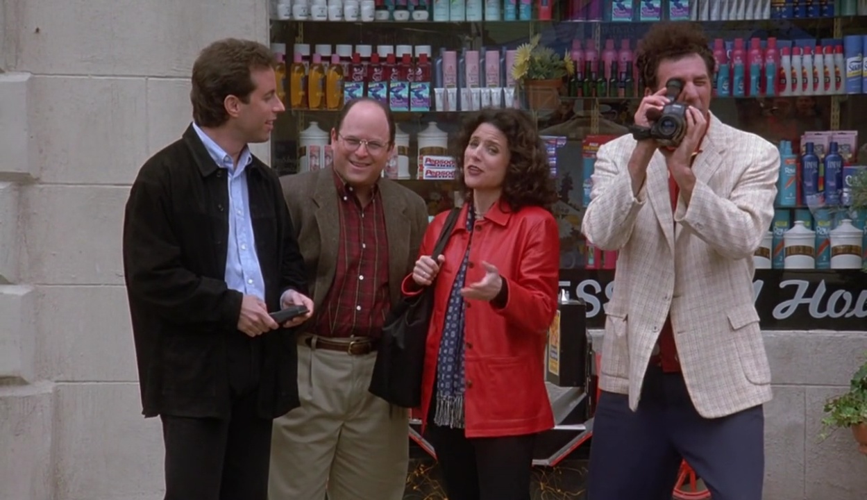 Four people are standing outside a store, with one holding a camcorder. The store display shows various personal care products.